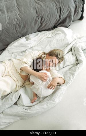 La petite fille s'allonge sur sa mère et veut dormir. Maman embrasse tendrement sa fille. Cadre vertical. Banque D'Images