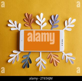 Un téléphone portable blanc avec un espace blanc orange vide pour le texte au milieu repose sur un fond jaune entouré de feuilles de papier de blanc, bleu et Banque D'Images