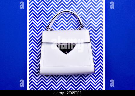 Sac moderne à poignée supérieure féminin en cuir blanc avec bordure noire, détail en métal et pieds isolés sur un fond bleu-blanc avec un motif zigzag. CRE Banque D'Images