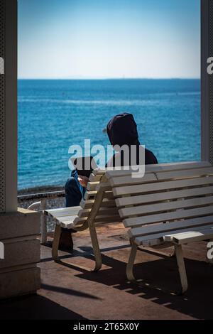 Guy dans un sweat à capuche assis sur un banc tout en travaillant à distance sur un ordinateur portable au Prom des Anglais, Nice, avec la mer en arrière-plan ; fond d'écran Remote Work Banque D'Images