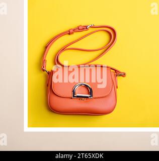 Sac à main pour femme en cuir orange vif tendance avec bandoulière et fermoir en métal isolé sur un fond géométrique jaune-beige-blanc. Mode essent Banque D'Images