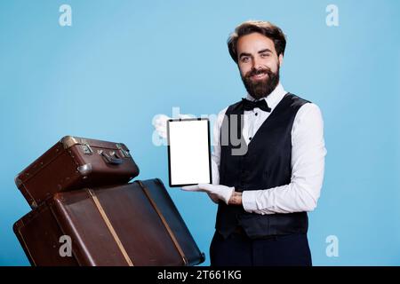 Bellboy présente un écran blanc sur tablette sur fond bleu, portant un costume formel et des gants à côté d'une pile de valises. Portier élégant affichant le modèle Copyspace sur l'appareil. Banque D'Images