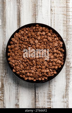 Gouttes de chocolat pour faire du chocolat, bonbons dans une assiette ronde noire sur un fond en bois clair. Concept de confiserie au chocolat tempéré, vue de dessus. Banque D'Images