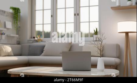 Un ordinateur portable sur une table basse dans un salon blanc moderne et confortable avec un canapé douillet contre la fenêtre, un lampadaire et des accessoires minimalistes Banque D'Images