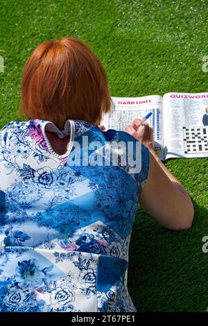 Femme avec des cheveux rouges / gingembre dans une robe à motifs bleus et roses couchée sur son devant sur de l'herbe artificielle au soleil faisant un livre de puzzle Banque D'Images