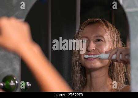 Jeune femme blonde se brosse les dents avec une brosse à dents électrique blanche dans la salle de bain regardant dans le miroir. L'hygiène, la notion de soins corporels. Banque D'Images