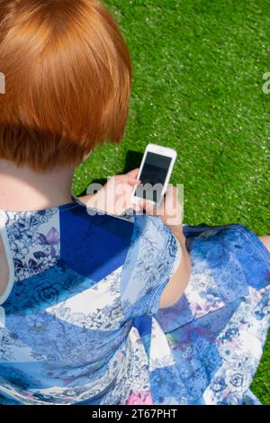 Une femme aux cheveux rouges portant une robe à motifs bleus, assise sur de l'herbe artificielle au soleil, regardant vers le bas et utilisant son iPhone Banque D'Images