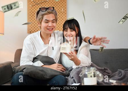 Heureux jeune couple tenant le modèle de maison assis sur le canapé avec des billets de dollar tombant d'en haut Banque D'Images