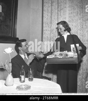 Dans les années 1950 Une fille de cigarette faisant le tour des tables dans le restautrant vendant des cigarettes de sa boîte vue devant elle. Un homme est vu recevant un paquet et a une pièce dans sa main à payer. Suède 1953 Kristoffersson ref BL39-12 Banque D'Images