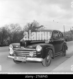 Dans les années 1940 Une jeune femme par sa voiture de tourisme de la marque automobile américaine Ford. Elle est vêtue d'un costume deux pièces, d'une jupe et d'une veste courte en tissu de laine à chevrons. Elle se tient sur les marches de la voiture pour atteindre le toit où un projecteur est monté. Il pourrait parfois être nécessaire de l'ajuster. Son chien peut être vu assis dans le siège du conducteur près du volant. La photo a été prise à Stockholm en Suède en 1949. Kristoffersson réf. AY83-6 Banque D'Images