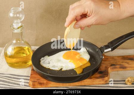 Femme main trempant du pain sur le jaune d'un œuf frit dans l'huile d'olive avec une poêle sur une planche de bois Banque D'Images