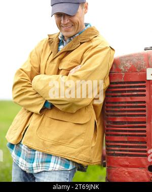 Vivre une vie bonne et simple. un fermier souriant debout à côté de son tracteur, les bras croisés et regardant vers le bas. Banque D'Images