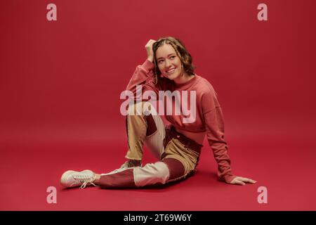 expression de soi, femme joyeuse en pantalon patchwork et manches longues court assis sur fond rouge Banque D'Images