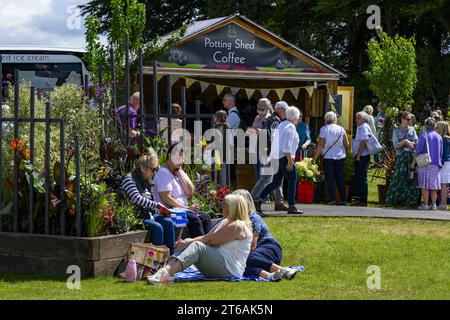 Visiteurs passant devant un stand de café et 4 quatre pique-niqueurs assis sur la pelouse en herbe au showground ensoleillé animé - 2023 Flower Show Tatton Park, Cheshire Angleterre Royaume-Uni. Banque D'Images