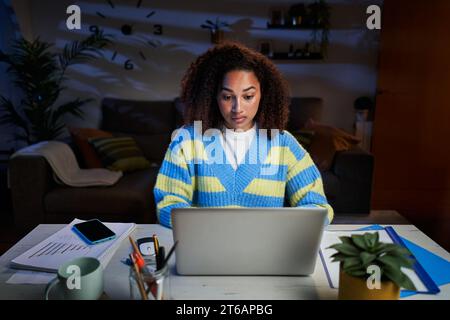 Jeune belle femme latine se concentrant à l'aide d'un ordinateur portable dans le salon la nuit. Étudiante afro. Banque D'Images