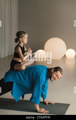 un enfant en position lotus est assis sur le dos d'un entraîneur de yoga pendant l'entraînement. Banque D'Images