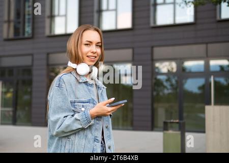 Jeune femme souriante avec des écouteurs sur le cou regarde dans la caméra tenant le téléphone. Joyeuse dame en veste denim textes meilleur ami après le travail pour traîner Banque D'Images