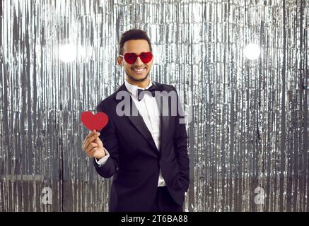 Heureux jeune homme noir dans des lunettes en forme de coeur, costume et noeud papillon à la fête de Saint-Valentin Banque D'Images