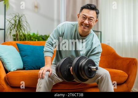 Haltères de levage de poids d'homme asiatique, biceps d'entraînement, entraînement, brûler les calories de graisse, entraînement de force pratiquant l'effort d'aérobic domestique de fitness à la maison appartement de chambre. Un homme adulte sportif est assis sur le canapé Banque D'Images