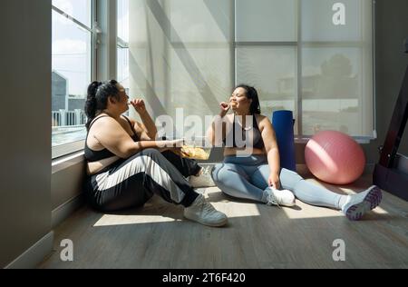 Deux femmes de grande taille appréciant joyeusement leur repas, assis dans une salle de fitness, favorisant la positivité corporelle et la vie joyeuse. Banque D'Images