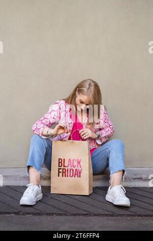 Femme blonde joyeuse est assise sur le steet tout en ouvrant un sac en papier à provisions avec un signe Black Friday Banque D'Images