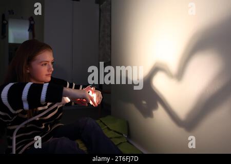 Adolescente faisant la forme de coeur d'ombre avec ses mains et doigts avec une lampe sur le mur Banque D'Images