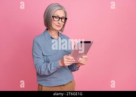 Photo de femme intelligente focalisée avec coiffure grise habillée chemise élégante bleue dans les lunettes lire les nouvelles sur tablette isolé sur fond rose Banque D'Images