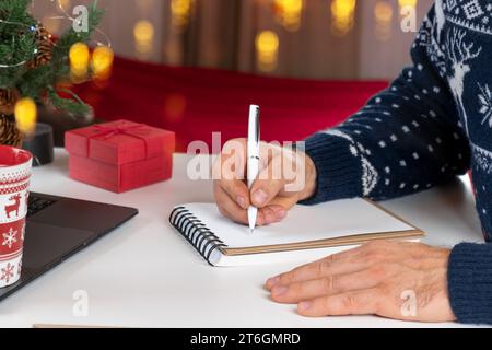 Ecriture d'une carte de vœux de Noël. Homme adulte faisant des cartes de voeux au bureau à domicile pendant Noël. Les mains de l'homme dans le chandail bleu tenant le papier vierge et envel Banque D'Images