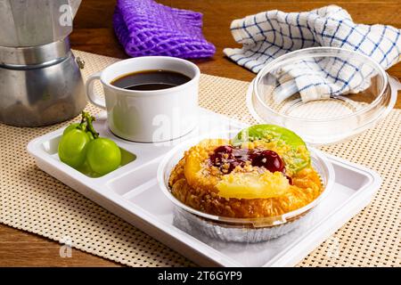 Vue à angle élevé de délicieux fruits mélangés maison danois, une tasse de café noir et des raisins dans un plateau en plastique blanc sur un tapis de table sur une table en bois. Banque D'Images