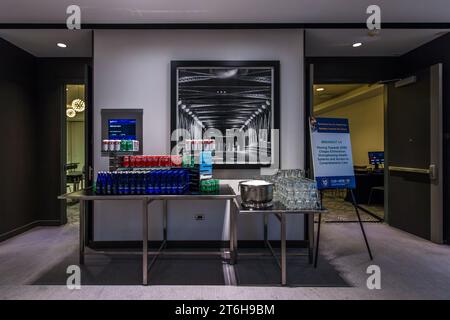 Une table de boissons avec des canettes et des bouteilles se trouve dans la zone de conférence du Radisson Blu Aqua Hotel à Chicago, Illinois, États-Unis, une grande photographie en noir et blanc d'un pont à treillis en acier est suspendue au-dessus, une pancarte à côté annonçant la session "Breakout 1.4 : Vers 2030 – Élimination de Chagas, Renforcement des Systèmes de Santé et Accès à des Soins Complets" d'une conférence internationale sur les maladies tropicales négligées. L'hôtel occupe les étages inférieurs de la tour Aqua, conçue en 2009 par l'architecte Jeanne Gang. Banque D'Images