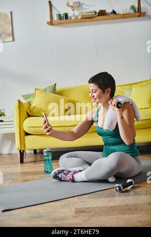 plan vertical de jeune femme heureuse assise sur le sol avec haltère et serviette prenant selfie, fitness Banque D'Images