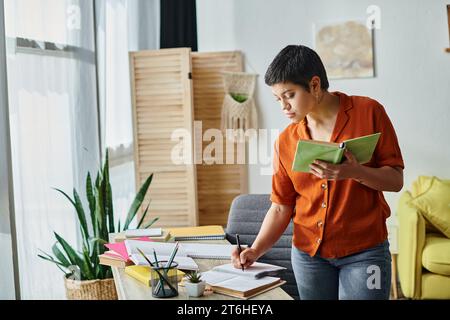 étudiant attirant concentré en chemise orange debout près du bureau tenant le livre prenant des notes, l'éducation Banque D'Images