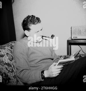 Années 1960, historique, un homme se relaxant, assis dans une chaise longue à motifs de fleurs fumant une pipe, lisant un livre, radio de l'époque à côté de lui, Angleterre, Royaume-Uni. Banque D'Images