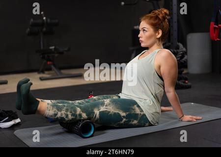 La femme s'engage dans un entraînement de récupération musculaire de base en utilisant un rouleau de mousse sur un tapis d'exercice dans la salle de gym. Banque D'Images