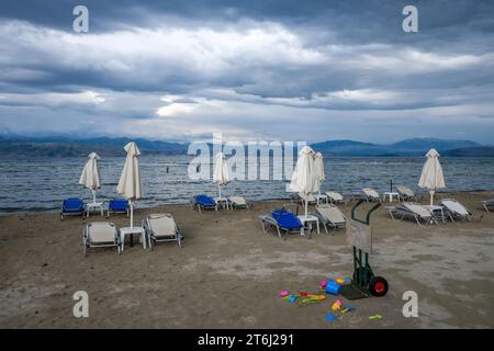 Kalamaki, Corfou, Grèce, chaises longues et parasols sur la plage de Kalamaki dans le nord-est de l'île grecque de Corfou, en arrière-plan le continent de l'Albanie avec la station balnéaire de Saranda. Devant les jouets oubliés pour enfants, seau et pelle. Location de parasols et chaises longues pour tableau de prix. Banque D'Images