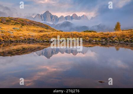 Italie, Vénétie, quartier de Belluno, Falcade, coucher de soleil brumeux en automne avec le côté nord de la Pale di San Martino reflété sur un étang alpin, Dolomites Banque D'Images