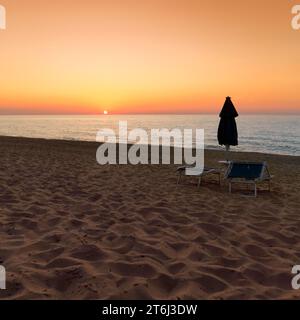 Coucher de soleil sur la mer, plage de sable déserte, transats abandonnés et parasol fermé, Spiaggia di Scivu, Arbus, Costa Verde, Sud Sardegna, Sardaigne, Italie Banque D'Images