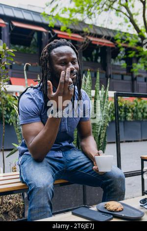 image verticale d'un jeune homme afro avec des dreadlocks, barbe et chemise, assis dehors parlant et se disputant avec quelqu'un et buvant du café, espace de copie Banque D'Images