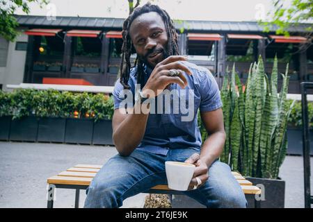 jeune homme africain avec barbe et dreadlocks heureux souriant, parlant et buvant du café assis sur un banc en bois à l'extérieur du café, espace de copie. Banque D'Images