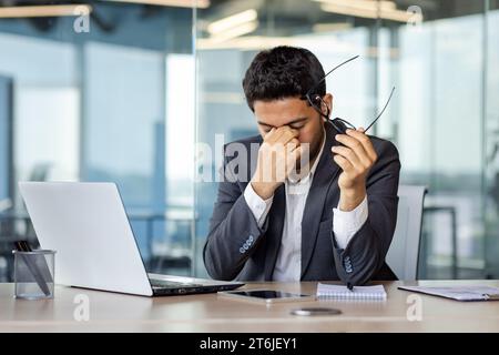 Homme d'affaires surfatigué et débordé au lieu de travail à l'intérieur du bureau, l'homme a enlevé les lunettes frottant les yeux, vertiges migraine et maux de tête, homme en costume d'affaires travaillant tard avec un ordinateur portable au lieu de travail. Banque D'Images