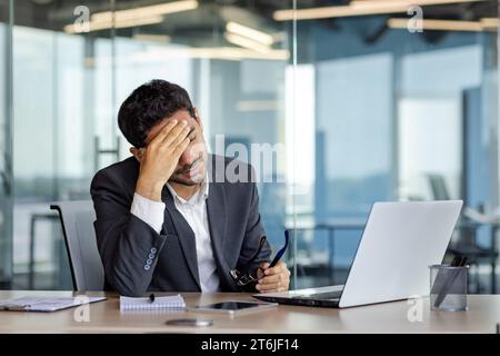 Homme d'affaires surfatigué et débordé au lieu de travail à l'intérieur du bureau, l'homme a enlevé les lunettes frottant les yeux, vertiges migraine et maux de tête, homme en costume d'affaires travaillant tard avec un ordinateur portable au lieu de travail. Banque D'Images