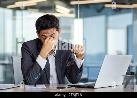 Homme d'affaires surfatigué et débordé au lieu de travail à l'intérieur du bureau, l'homme a enlevé les lunettes frottant les yeux, vertiges migraine et maux de tête, homme en costume d'affaires travaillant tard avec un ordinateur portable au lieu de travail. Banque D'Images