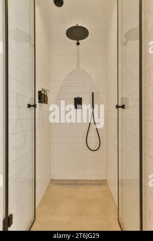 une douche de plain-pied qui est blanche et a des luminaires noirs sur le mur, avec une porte de douche ouverte Banque D'Images