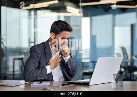 Homme d'affaires malade toussant à l'intérieur du bureau au lieu de travail, homme en costume d'affaires travaillant avec un ordinateur portable, rhume avec la grippe. Banque D'Images