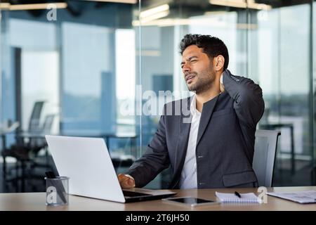 Douleur au cou, homme surmené et overfatigué travaillant tard, homme d'affaires massant le muscle du cou, travaillant assis sur une chaise à la table avec un ordinateur portable à l'intérieur du bureau au lieu de travail. Banque D'Images