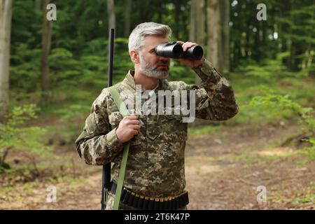 Homme avec un fusil de chasse regardant à travers des jumelles dans la forêt Banque D'Images