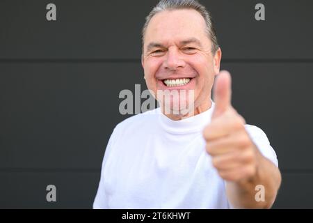 Un homme mûr souriant montrant ses nouvelles dents et donnant un geste pouce levé devant un mur noir Banque D'Images