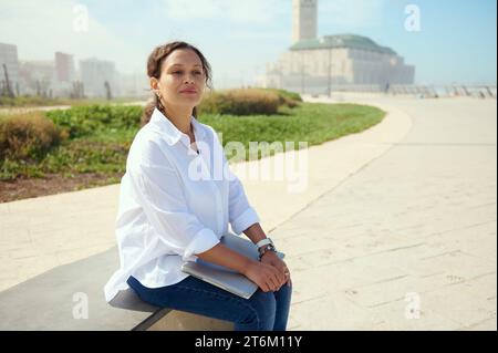 Femme d'affaires confiante, entrepreneur indépendant assis sur un banc à l'extérieur contre un fond de mosquée Hassan II, pensant à un nouveau projet de start-up, dre Banque D'Images