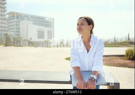 Portrait confiant authentique de jeune adulte multi-ethnique femme, femme d'affaires, freelance, entrepreneur, développeur assis sur un banc contre cityscap Banque D'Images