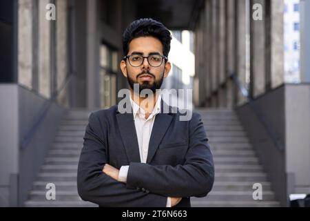 Portrait d'un homme d'affaires hindou sérieux debout à l'extérieur du bureau avec ses bras pliés sur sa poitrine, un patron strict dans des lunettes et un costume regardant avec confiance dans la caméra. Banque D'Images